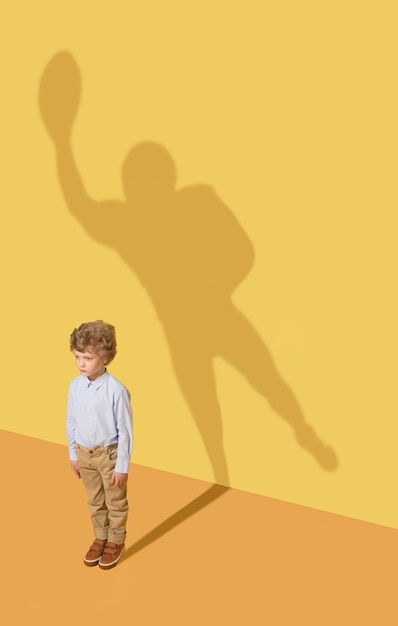 Foto gratuita mejor miembro del equipo. concepto de infancia y sueño. imagen conceptual con niño y sombra en la pared amarilla del estudio. el niño pequeño quiere convertirse en jugador de fútbol americano y construir una carrera deportiva.