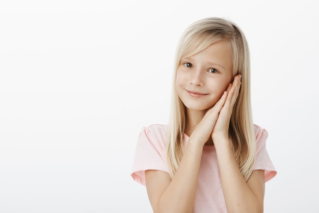 El mejor momento para pasar es dormir. Retrato de una niña adorable de aspecto amigable con cabello rubio, sonriendo alegremente y sosteniendo las palmas juntas cerca de la mejilla, de pie como un ángel sobre una pared gris