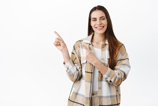 La mejor oferta en el mercado Hermosa mujer sonriente señalando con el dedo a un lado mostrando un anuncio en el lado izquierdo de la pantalla de información de copyspace de pie sobre fondo blanco