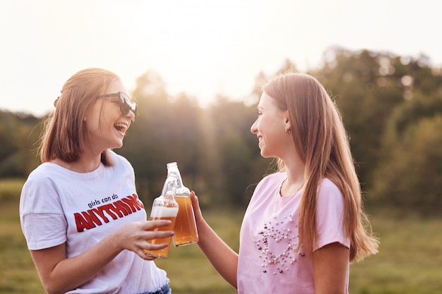 Las mejores amigas brindan con botellas de cerveza fría, se divierten juntas, pasan tiempo libre al aire libre