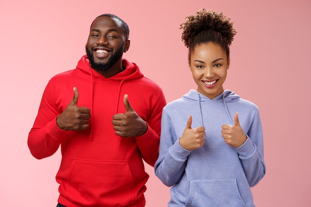 Los mejores amigos apoyan tu elección. Retrato orgulloso satisfecho apuesto hombre afroamericano mujer mostrar cámara pulgar hacia arriba sonriendo ampliamente animando gusta compra impresionante, de pie fondo rosa