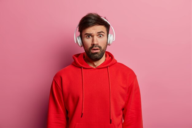 Meloman masculino estupefacto mira sorprendentemente, escucha audio a través de auriculares, vestido con una sudadera roja, escucha noticias asombrosas, posa sobre una pared rosada. Gente, reacción, emociones.