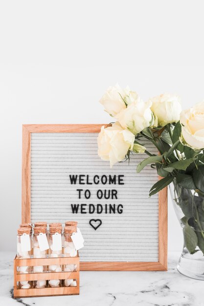 Mensaje de bienvenida para bodas en marco blanco con tubos de ensayo de malvavisco y florero de rosas