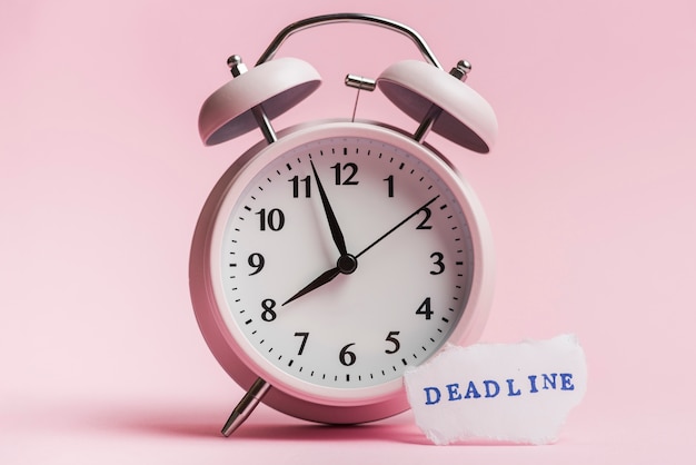 Mensaje de fecha límite en papel rasgado cerca del reloj de alarma con fondo rosa