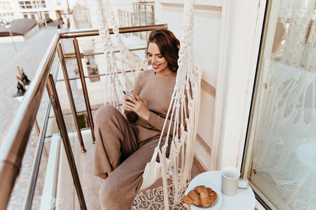 Mensaje de texto de chica agradable durante el almuerzo en el balcón. Elegante mujer joven sentada en la terraza con croissant y té.