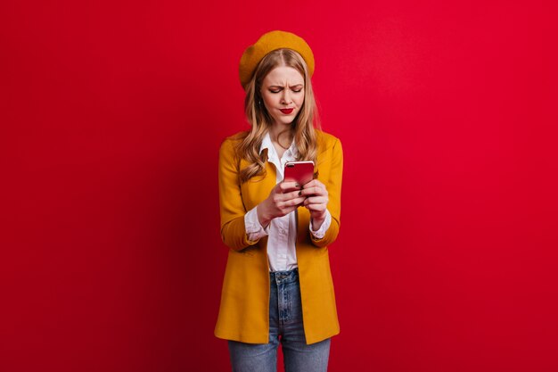 Mensaje de texto de chica francesa concentrada. Mujer joven rubia en ropa casual con smartphone en pared roja.