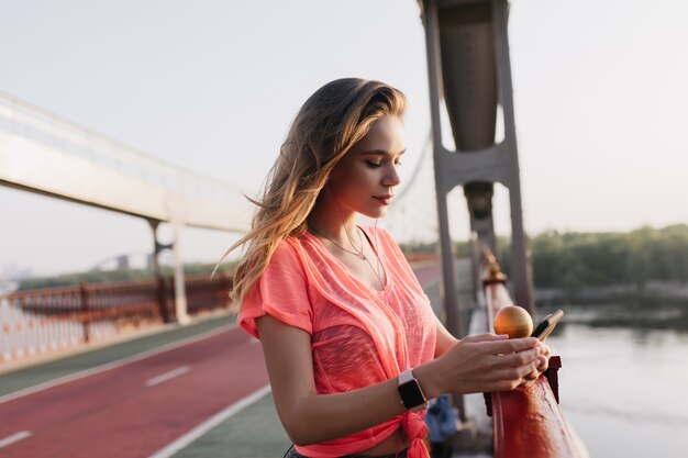 Mensaje de texto de chica rubia pensativa mientras está de pie cerca del camino de ceniza. Hermosa mujer en ropa casual posando al aire libre después del entrenamiento.