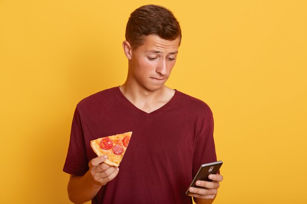 Mensaje de texto joven en teléfono inteligente aislado sobre la pared amarilla. Hombre pensativo con teléfono móvil y una porción de pizza en las manos