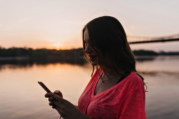 Mensaje de texto de linda chica con sonrisa en la naturaleza. Encantadora mujer europea posando con teléfono cerca del río.