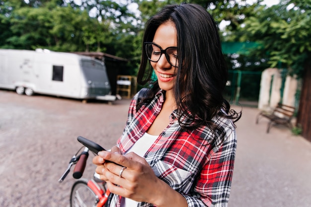 Mensaje de texto de mujer morena elegante en la calle. Chica de buen humor en elegante camisa mirando la pantalla del teléfono con una sonrisa feliz.