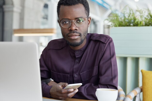 Mensaje de tipos de trabajador corporativo serio confiado en un teléfono inteligente, vestido formalmente, se sienta frente a una computadora portátil genérica