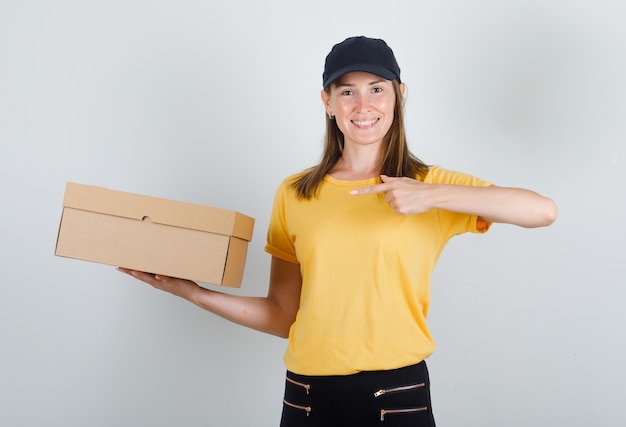 Mensajero femenino dedo acusador en caja de cartón en camiseta, pantalón, gorra y mirando contento