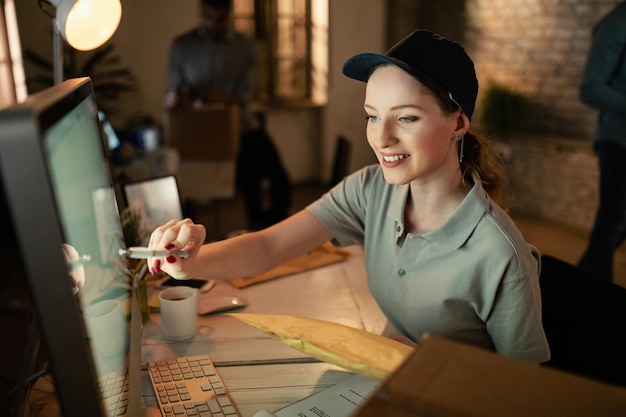 Mensajero femenino feliz que usa la computadora y lee datos para la entrega de paquetes mientras trabaja en la oficina