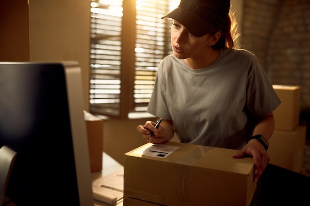 Mensajero femenino joven que usa la computadora mientras prepara el paquete para una entrega en la oficina