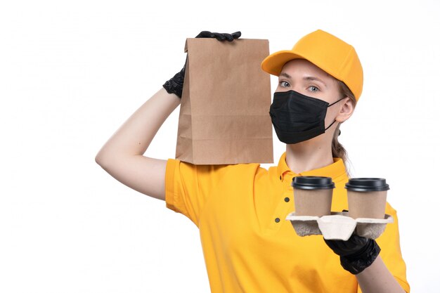 Un mensajero femenino joven de la vista frontal en guantes negros uniformes amarillos y máscara negra que sostiene el paquete de comida y las tazas de café