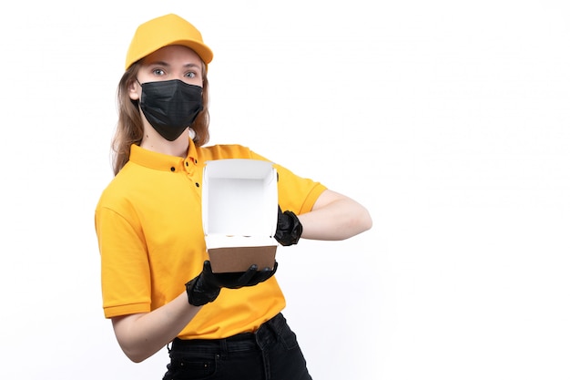 Un mensajero femenino joven de la vista frontal en guantes negros uniformes amarillos y máscara negra que sostiene un paquete de comida vacío