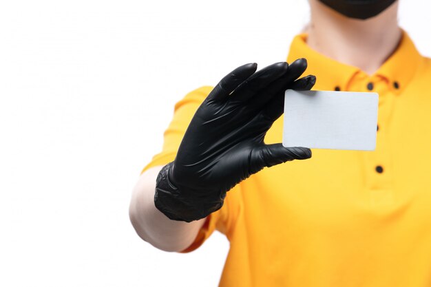 Un mensajero femenino joven de la vista frontal en guantes negros uniformes amarillos y máscara negra que sostiene la tarjeta blanca