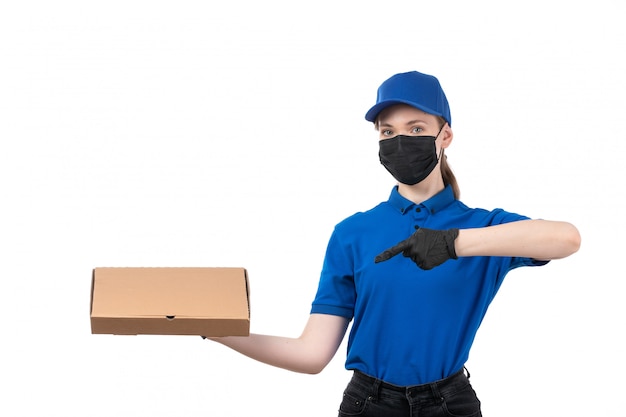 Un mensajero femenino joven de la vista frontal en guantes negros uniformes azules y máscara negra que sostiene el paquete de entrega de alimentos