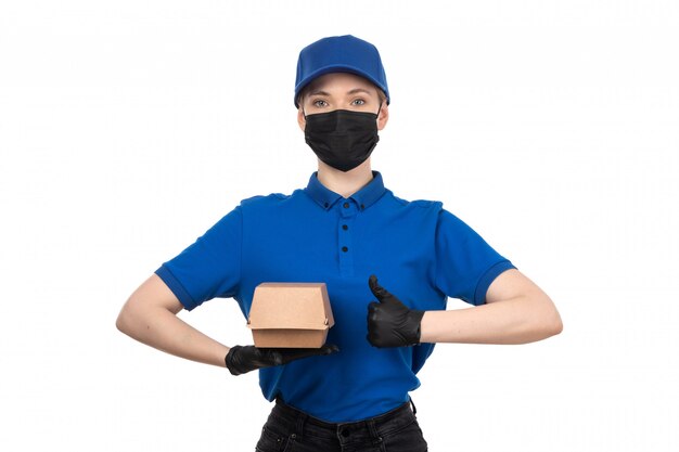 Un mensajero femenino joven de la vista frontal en máscara negra uniforme azul y guantes que sostienen el paquete de entrega de alimentos