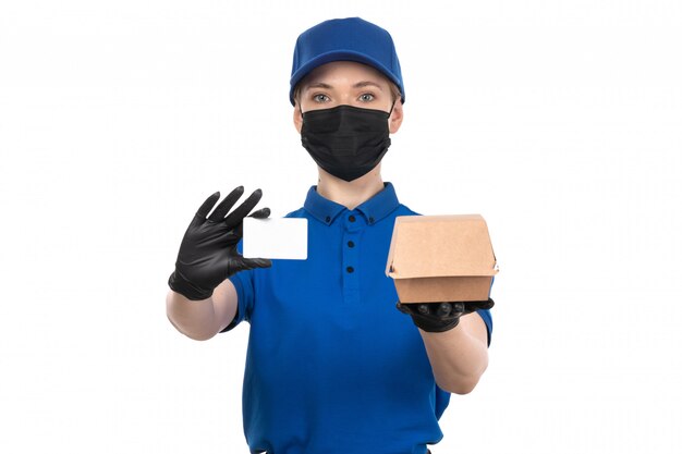 Un mensajero femenino joven de la vista frontal en máscara negra uniforme azul y guantes que sostienen el paquete de entrega de alimentos