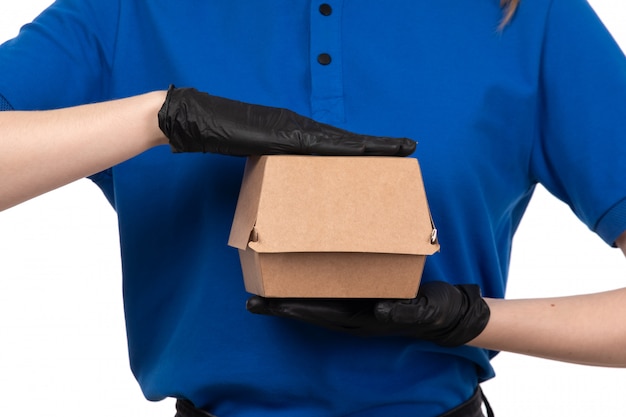 Un mensajero femenino joven de la vista frontal en máscara negra uniforme azul y guantes que sostienen el paquete de entrega de alimentos