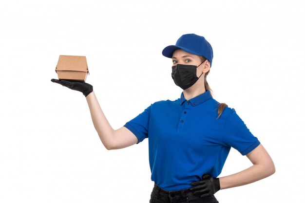 Un mensajero femenino joven de la vista frontal en máscara negra uniforme azul y guantes que sostienen el paquete de entrega de alimentos