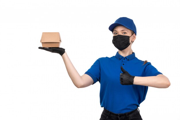 Un mensajero femenino joven de la vista frontal en máscara negra uniforme azul y guantes que sostienen el paquete de entrega de alimentos