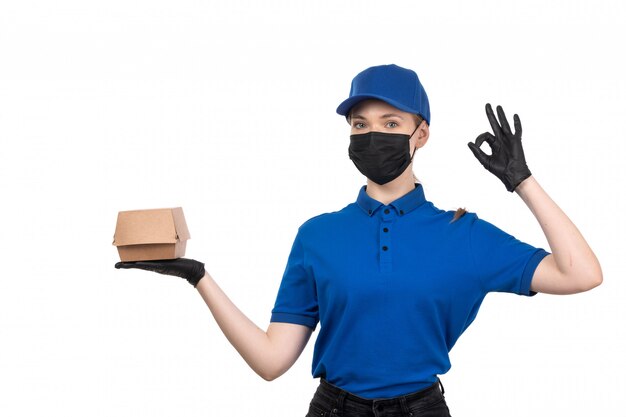 Un mensajero femenino joven de la vista frontal en máscara negra uniforme azul y guantes que sostienen el paquete de entrega de alimentos