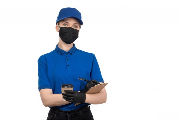 Un mensajero femenino joven de la vista frontal en máscara negra uniforme azul y guantes que sostienen la taza de café y el bloc de notas