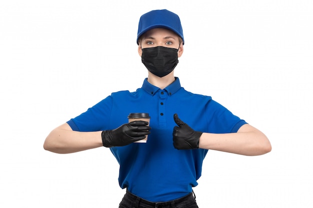 Un mensajero femenino joven de la vista frontal en máscara negra uniforme azul y guantes que sostienen la taza de café