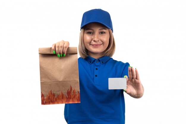 Un mensajero femenino joven de la vista frontal en uniforme azul que sostiene el paquete de entrega de alimentos y la tarjeta blanca