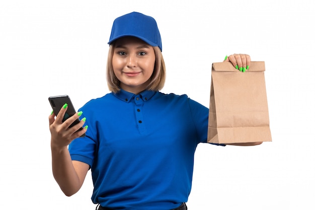 Un mensajero femenino joven de la vista frontal en uniforme azul que sostiene el teléfono y el paquete de entrega de alimentos