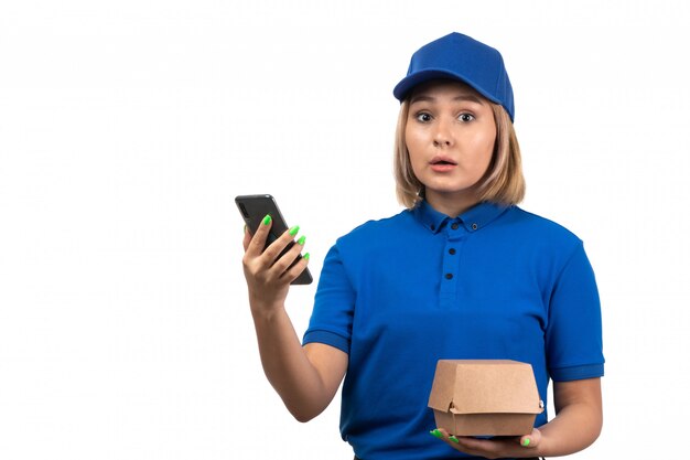 Un mensajero femenino joven de la vista frontal en uniforme azul que sostiene el teléfono y el paquete de entrega de alimentos