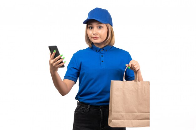 Un mensajero femenino joven de la vista frontal en uniforme azul que sostiene el teléfono y el paquete de entrega de alimentos