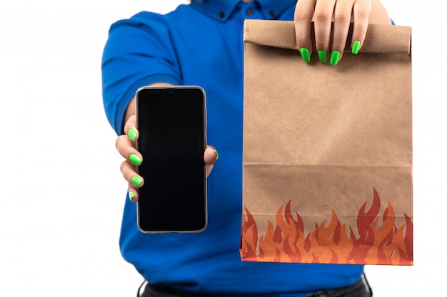 Un mensajero femenino joven de la vista frontal en uniforme azul que sostiene el teléfono y el paquete de entrega de alimentos