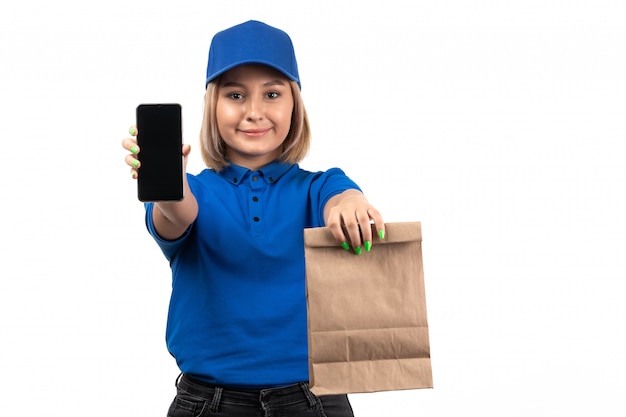 Un mensajero femenino joven de la vista frontal en uniforme azul que sostiene el teléfono y el paquete de entrega de alimentos