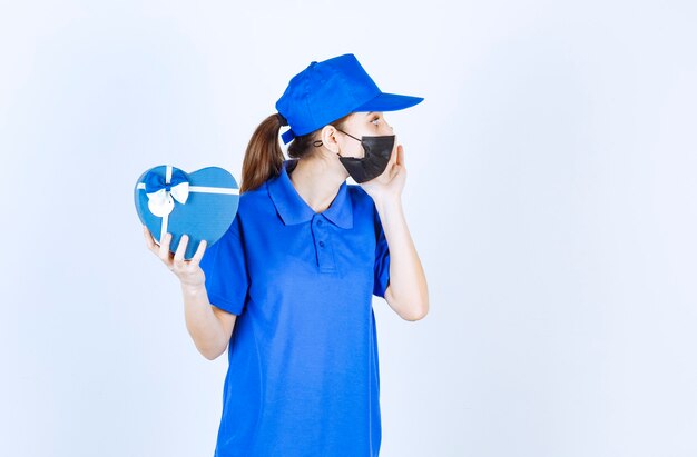 Mensajero femenino en máscara y uniforme azul sosteniendo una caja de regalo con forma de corazón y llamando a alguien para recibirlo