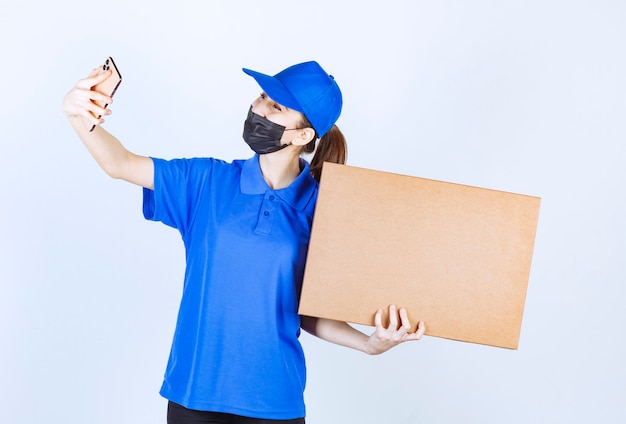 Mensajero femenino con máscara y uniforme azul sosteniendo un gran paquete de cartón y tomando nuevos pedidos en su teléfono