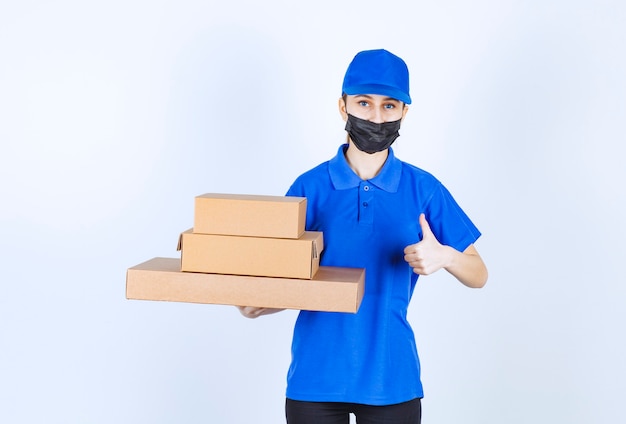 Mensajero femenino en máscara y uniforme azul sosteniendo un stock de cajas de cartón y mostrando un signo de mano positivo