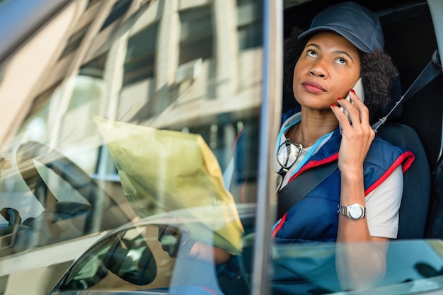 Mensajero femenino negro que se comunica por teléfono móvil mientras entrega paquetes con una furgoneta