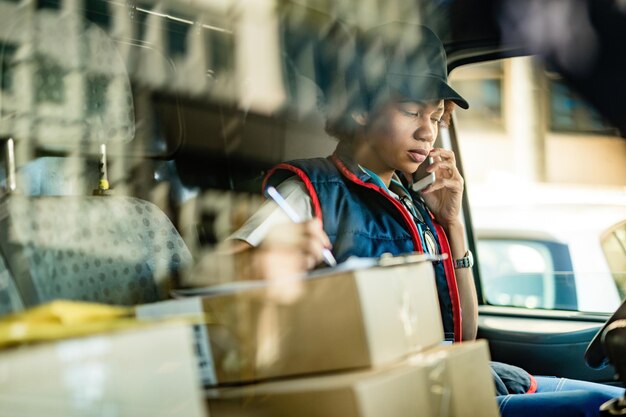 Mensajero femenino negro que se comunica por teléfono móvil y toma notas mientras está sentado en la camioneta de reparto La vista es a través de la ventana