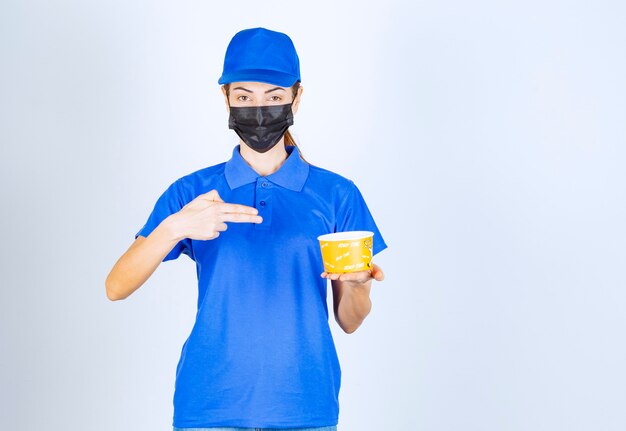 Mensajero femenino del restaurante en uniforme azul y mascarilla sosteniendo una comida para llevar.