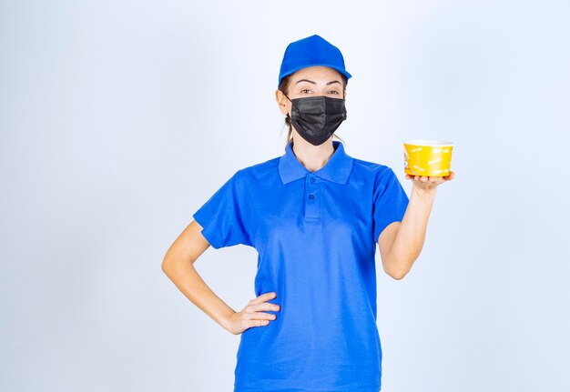 Mensajero femenino del restaurante en uniforme azul y mascarilla sosteniendo una comida para llevar.