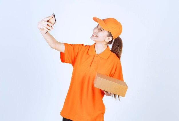 Mensajero femenino en uniforme amarillo entregando una caja de cartón y haciendo una videollamada al cliente