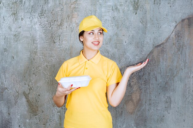 Mensajero femenino en uniforme amarillo entregando una caja de comida para llevar blanca y apuntando al cliente