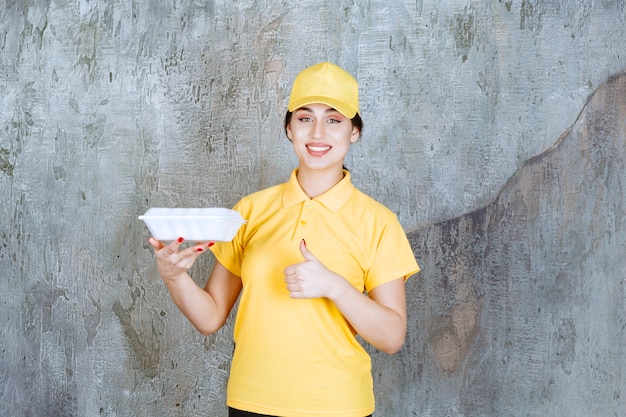 Mensajero femenino en uniforme amarillo entregando una caja de comida para llevar blanca y disfrutando de la comida