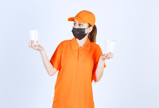 Mensajero femenino en uniforme amarillo y máscara negra sosteniendo bebidas para llevar en vasos de plástico en ambas manos