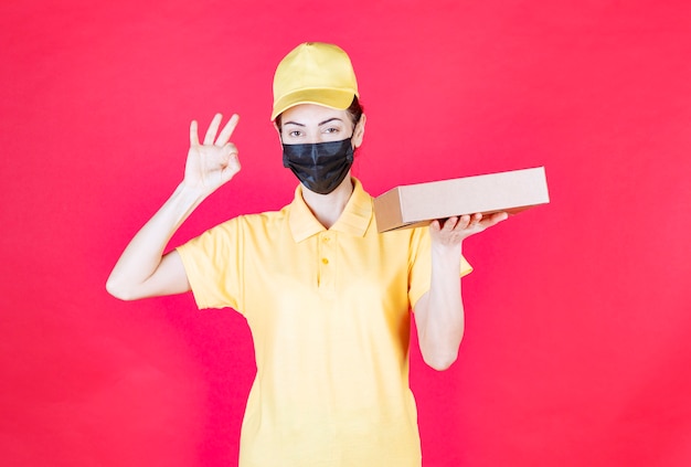 Mensajero femenino en uniforme amarillo y máscara negra sosteniendo la caja de cartón y mostrando el signo de disfrute