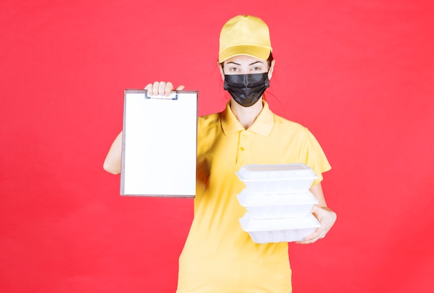 Mensajero femenino en uniforme amarillo y máscara negra sosteniendo múltiples paquetes para llevar y presentando la lista de firmas