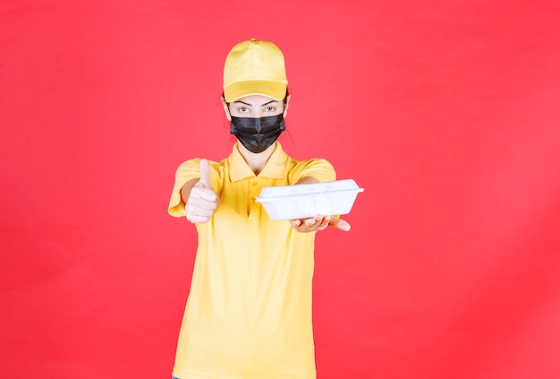 Foto gratuita mensajero femenino en uniforme amarillo y máscara negra sosteniendo un paquete de comida para llevar y mostrando un signo de mano positivo